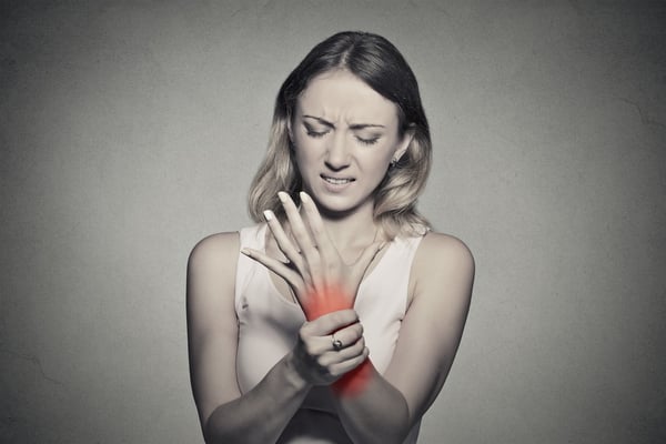 Young woman holding her painful wrist isolated on gray wall background. Sprain pain location indicated by red spot. Negative face expression